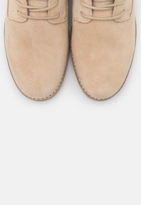 Chaussures en daim beige à bouts arrondis, surface texturée et lacets marron clair. Semelles en caoutchouc et coutures apparentes le long du bord.