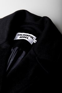 Gola de um casaco preto de lã com etiqueta da marca "Colourful Rebel" e etiqueta de tamanho S dentro do decote, contra um fundo claro.