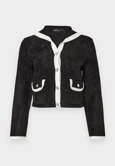 Schwarze, kurz geschnittene Jacke aus weichem Stoff mit einem weißen Matrosenkragen, silbernen Knöpfen und zwei vorderen Taschen mit weißem Besatz.