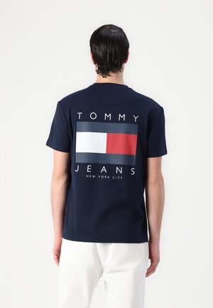 Mężczyzna ubrany w granatowy t-shirt Tommy Jeans z czerwono-biało-niebieskim logo oraz białe spodnie, widoczny od tyłu na jednolitym tle.