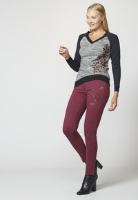 Grau-und-schwarzer langärmeliger V-Ausschnitt-Pullover mit floraler Stickerei, kombiniert mit burgunderroten Skinny-Jeans mit Akzentdetails und schwarzen High-Heels.