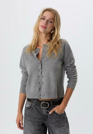 Frau mit langen blonden Haaren, die eine geknöpfte graue Strickjacke, einen schwarzen Gürtel und ausgebleichte schwarze Jeans trägt, mit einer Hand in der Tasche vor einem einfarbigen Hintergrund.