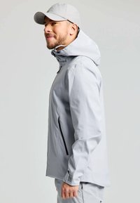 Giacca impermeabile grigia chiara con cappuccio, caratterizzata da dettagli in zip nera e polsini regolabili. Texture liscia, vestibilità rilassata, design snello.