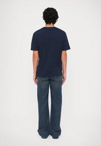 Hombre con cabello rizado vistiendo una camiseta básica azul marino y unos pantalones vaqueros oscuros holgados, de pie mirando hacia atrás contra un fondo liso.