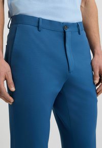 Blauwe op maat gemaakte broek met een gladde textuur, voorzien van een ritssluiting, knoopsluiting en zijzakken. De tailleband heeft riemlussen.