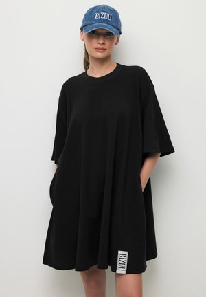 Kobieta w luźnej czarnej sukience z krótkimi rękawami i niebieskiej czapce z białym logo „BIZUU”, stojąca na tle gładkiego jasnego tła.