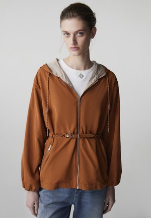 Młoda kobieta w rudobrązowej kurtce z kapturem zapinanej na zamek, z paskiem, na białej koszuli i niebieskich dżinsach, stojąca na tle jednolitego jasnego tła.