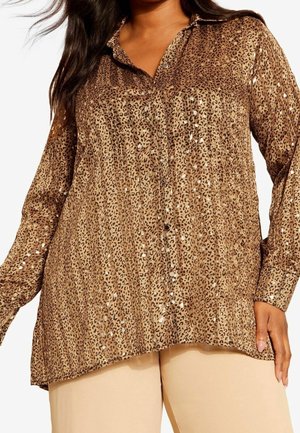 Kobieta ubrana w brązową bluzkę na guziki z nadrukiem lamparta i długimi rękawami oraz beżowe spodnie, pokazana od ramion do bioder.