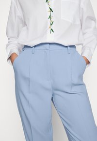 Tailored light blue trousers with a smooth texture, featuring front pleats and side pockets. Paired with a white shirt.