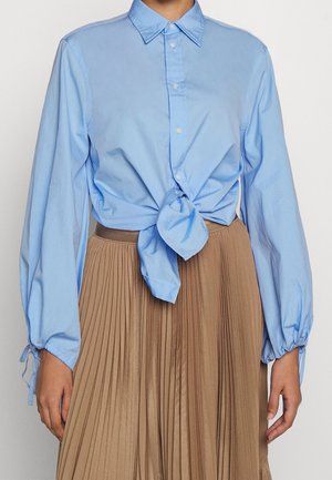 Blauw overhemd met lange mouwen en knopen, vooraan in een knoop gebonden, gecombineerd met een hoog getailleerde, geplisseerde beige rok.