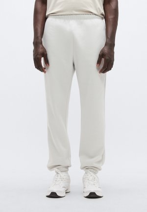 Gaiši pelēkas sporta bikses ar elastīgu jostu un aprocēm, izgatavotas no mīksta materiāla. Attēlota persona baltās sporta kurpēs.