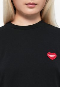 Black cotton t-shirt with a round neckline and short sleeves. Features a red heart-shaped embroidered logo on the chest.