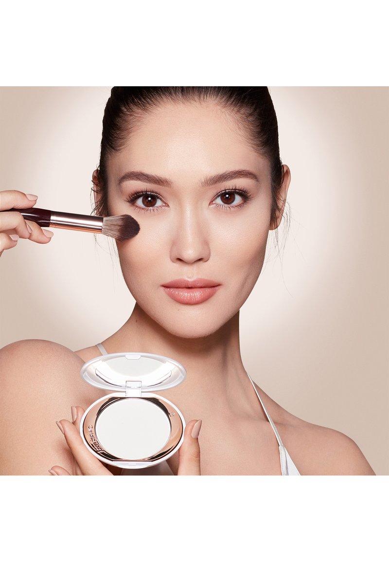 Woman applies face powder on cheek with brush while holding a compact mirror powder case, neutral beige background.
