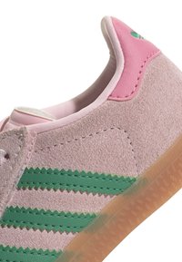 Pink suede sneaker featuring green zigzag stripes, a pink leather heel tab, and a translucent rubber sole with texture patterns.