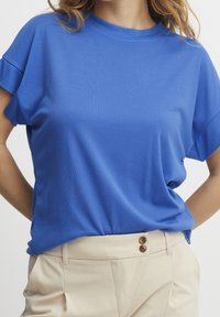 Donna che indossa una blusa corta blu royal a maniche corte, sblusata dentro pantaloni beige a vita alta con doppio bottone.