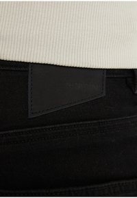 Tela vaquera negra con una cintura visible en crema con acanaladuras. Presenta una etiqueta de cuero negro con texto en relieve. Costuras limpias en toda la prenda.