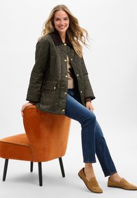 Olijfgroen gewatteerde jas met een bruine corduroy kraag, gouden drukknoopjes, twee voorkuiden, gecombineerd met blauwe jeans en tan instapschoenen.
