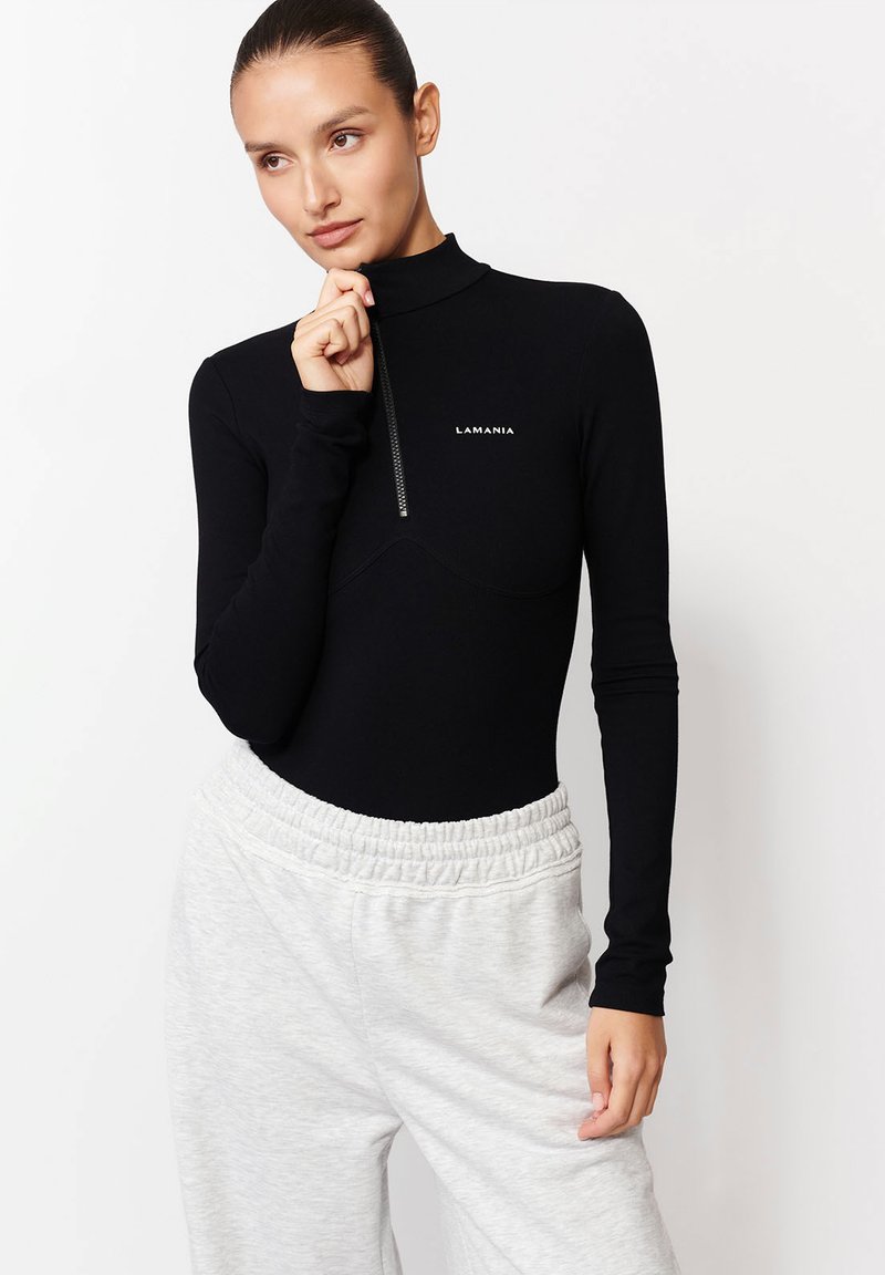 Žena oblečená v čiernom tričku s dlhým rukávom a krátkym zipsom so značkovým logom "LAMANIA", skombinovaná so svetlo šedými nohavicami s elastickým pásom.