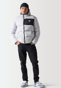 Chaqueta de plumas en gris claro con un panel negro en el pecho, cierre con cremallera y capucha; combinada con jeans oscuros y zapatillas azules.