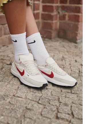 Jambes portant des chaussettes blanches Nike et des baskets blanches Nike avec un swoosh rouge, debout sur un trottoir pavé près d'un mur en briques.