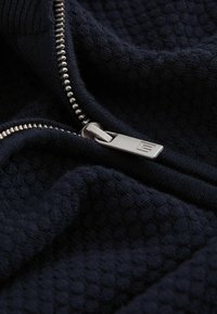 Pulover cu fermoar de culoare bleumarin, realizat dintr-un material texturat și cu fermoar argintiu. Prim-planul arată detaliile fermoarului și ale textilelor moi.
