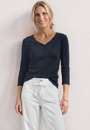 Kobieta z blond włosami związanymi do tyłu, ubrana w ciemną bluzkę z długim rękawem i białe spodnie, stojąca przy jednolitej jasnej ścianie, uśmiechająca się.