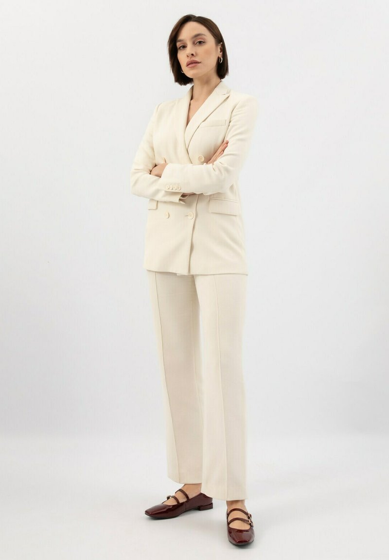 Kremowa marynarka o podwójnym zapięciu, dopasowane spodnie, oraz burgundowe buty na paski. Gładka tekstura, minimalistyczny styl.