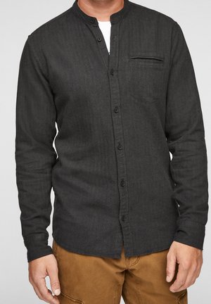 Mann trägt ein schwarzes Hemd mit Stehkragen über einem weißen Unterhemd und braunen Hosen, steht vor einem grauen Hintergrund.