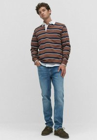 Brown striped long-sleeve top over a white collared shirt, paired with light blue jeans and brown shoes. Neutral background.