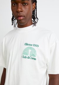 Hvitt bomull t-skjorte med grønn brodert tekst: "Ellesse 1959" og et geometrisk design med "Club di Corsa" under, rund hals-stil.