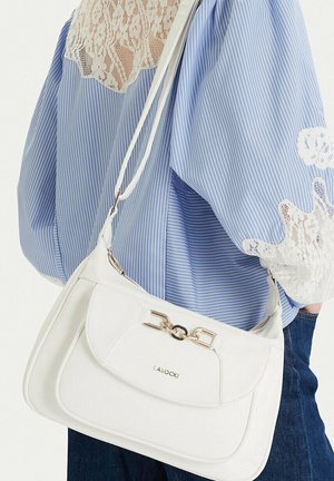 Bílá kožená taška přes rameno se zlatou sponou nošená přes rameno, doplněná modrou pruhovanou blůzou s krajkovými detaily a tmavými džínami.