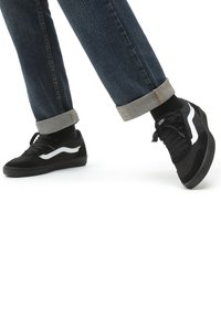 Black sneakers with white accents, featuring a mix of suede and mesh materials. Worn with cuffed dark jeans and black socks.