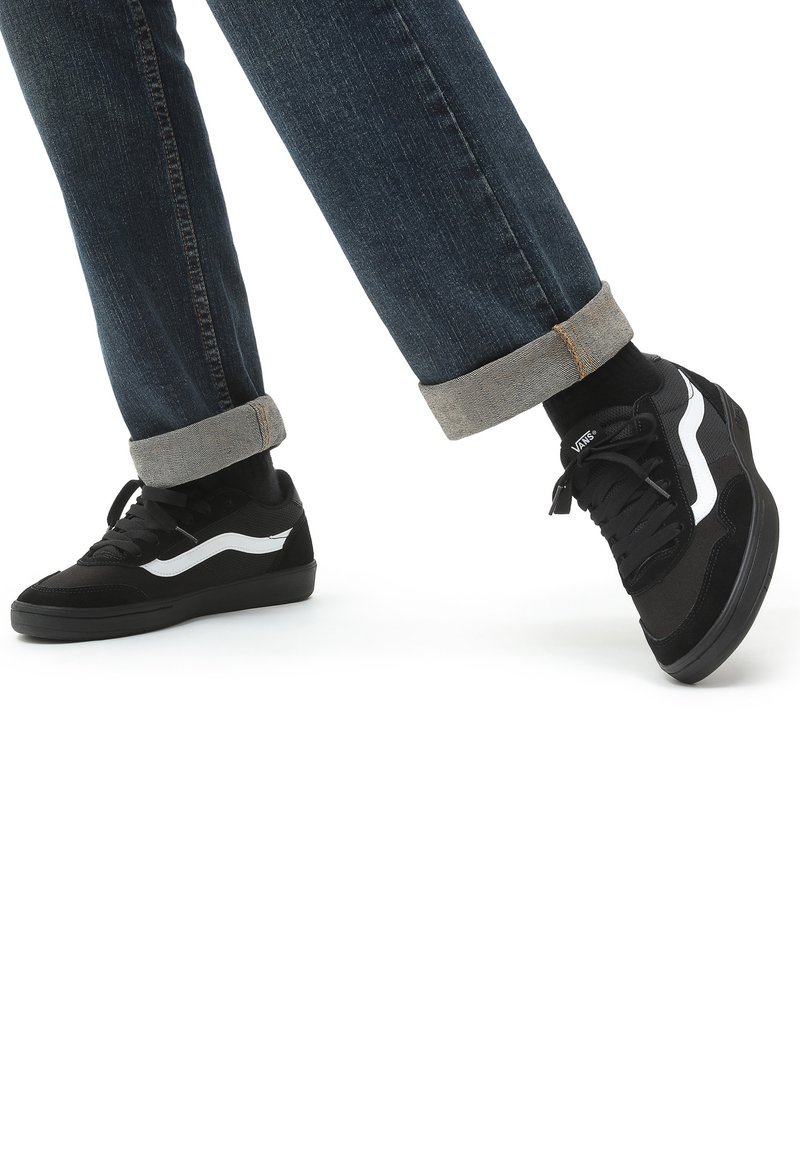 Black sneakers with white accents, featuring a mix of suede and mesh materials. Worn with cuffed dark jeans and black socks.