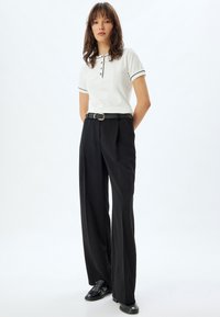 Camisa polo blanca con ribete negro y tapeta de botones, combinada con pantalones de pierna ancha en negro de tiro alto y zapatos de cuero negros.