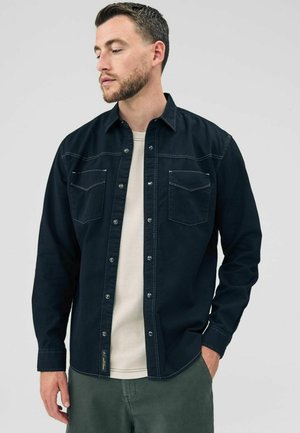 Dark blue denim shirt with snap buttons, two chest pockets, and contrast stitching, worn over a light t-shirt and green pants.