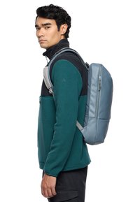 Un sac à dos gris rembourré au design épuré, avec une poche zippée à l'avant, porté sur une veste en polaire verte. Bretelles réglables pour plus de confort.