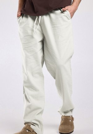 Uomo che indossa pantaloni larghi chiari con coulisse, mani in tasca e scarpe slip-on in camoscio marrone su sfondo semplice.