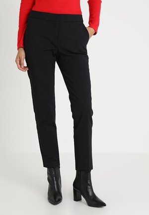 Femme portant un pantalon noir slim avec poches latérales, associé à des bottines noires à talons et un haut rouge à manches longues, debout avec une main dans la poche.