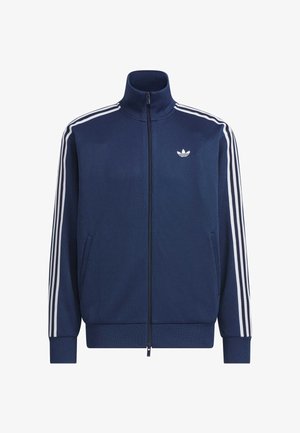 Chaqueta deportiva con cremallera azul marino, con tres rayas blancas en las mangas y el logo blanco de Adidas en el pecho, puños y cintura acanalados.