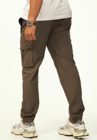 Braune Cargo-Hosen aus weichem Stoff, mit Seitentaschen, elastischen Bündchen und Kordelzugdetails, kombiniert mit grau-weißen Sneakers.