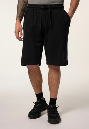 Mand iført sorte snørebåndsshorts, sorte sokker og sorte træningssko, stående med den ene hånd i lommen mod en ensfarvet baggrund.