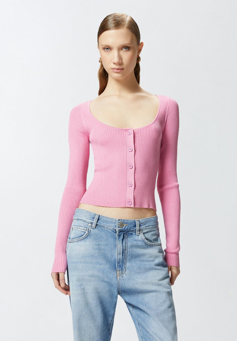 Ružičasti rebrasti kardigan dugih rukava s izrezom oko vrata i prednjim dugmadi, uparen s svijetloplavim trapericama.