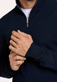 Pullover a coste navy con colletto alto e zip argentata, dotato di polsini aderenti e texture morbida. Mani che afferrano il tessuto.
