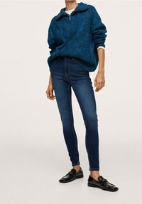 Suéter texturizado en color azul verdoso con cremallera de un cuarto, combinado con jeans ajustados de un azul oscuro. Mocasines negros completan el conjunto.