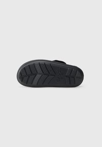 Crni slip-on papuč sa soleom s uzorkom i vidljivim imenom brenda "Crocs" na jednobojnoj svijetloj pozadini.