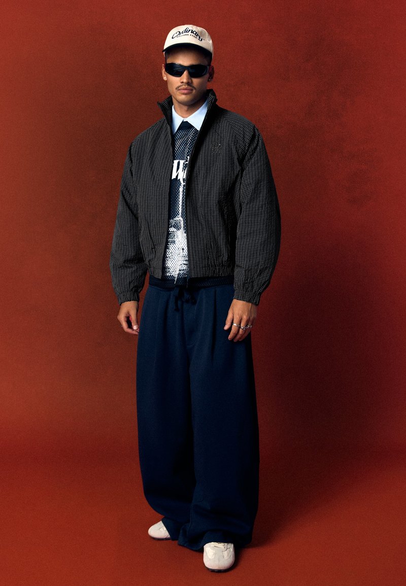 Homme portant une veste à carreaux noire, un t-shirt graphique, un pantalon large bleu marine, des baskets blanches, des lunettes de soleil noires et une casquette blanche, debout devant un fond marron.