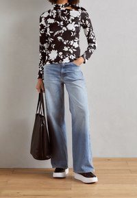 Zwarte bloementop met lange mouwen en witte print, gecombineerd met lichtblauwe wijde jeans en plateauschoenen. Een brown tote bag hangt aan één hand.