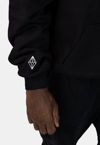 Zwarte sweatshirt met geribbelde manchetten en een geborduurd wit diamantlogo op de mouw. De stof lijkt zacht en gestructureerd.