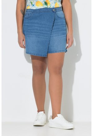 Frau trägt einen blauen Jeans-Wickelrock, weiße Sneakers und ein Top mit Zitronenmuster, steht vor weißem Hintergrund.