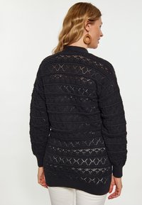 Maglione nero lavorato a maglia con un design a rete e motivi testurizzati, rifinito con polsini a coste. Maniche lunghe e orlo posteriore leggermente più lungo.
